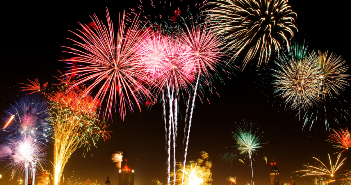 New Year’s Eve fireworks illuminate a crowded city square filled with cheering people and festive lights