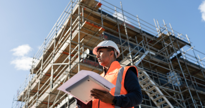 A construction worker with safety gear at a German construction site holding blueprints.